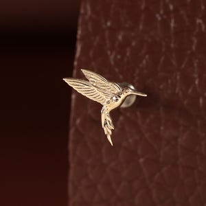 May include: A small gold-toned stud earring shaped like a hummingbird in flight. The metal piece features engraved wing and tail feather details, with a pointed beak extending forward and a post backing visible behind the bird figure.