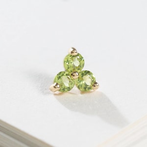 May include: A small stud earring featuring three round, light green peridot gemstones arranged in a triangular cluster. The stones are set in a gold-toned metal setting with small prongs securing each individual gemstone in place.