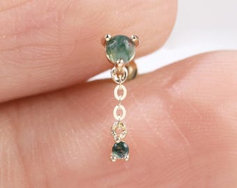 May include: A gold-colored earring with three green gemstones. The top gemstone is round, followed by a chain of small gold links, and a smaller, teardrop-shaped green gemstone at the bottom. The earring is held by a finger.
