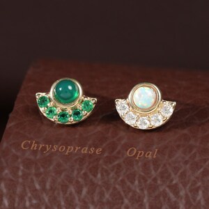 May include: Two gold stud earrings shaped like semi-circles. The left earring features a central green chrysoprase cabochon surrounded by small green stones. The right earring features a central white opal cabochon surrounded by small clear stones.