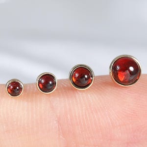 May include: Four round stud earrings featuring deep red garnet gemstones set in polished gold-toned metal bezels. The earrings are arranged in a row by size, from smallest to largest, showcasing a smooth, cabochon-style finish on each stone.