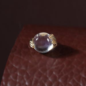 May include: A gold ring featuring a round, translucent, light purple cabochon gemstone. The metal band has a textured, ribbed design that wraps around the base of the stone, creating a detailed setting for the smooth, polished, spherical gem.