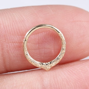 May include: A gold-colored, hinged nose ring with an ornate, engraved design. The ring features a decorative, pointed bottom and detailed scrollwork along the sides. The jewelry is held between two fingers against a neutral background.