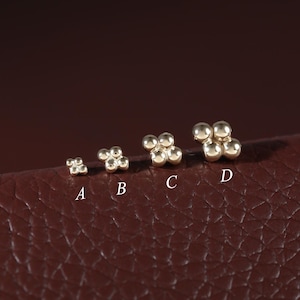 Pode incluir: Quatro brincos de pino em tom dourado dispostos em fila, rotulados A, B, C e D. Cada brinco apresenta um grupo de quatro pequenas contas esféricas dispostas em formação quadrada, com o tamanho dos grupos aumentando progressivamente de A a D.