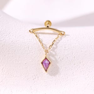 May include: A gold earring featuring a small stud and a fine chain. The chain supports a purple, diamond-shaped gemstone. The earring is set against a white, textured backdrop.