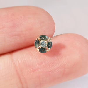 May include: A small, round stud earring with a gold setting. The earring features a cluster of small, dark green and light blue gemstones arranged in a square pattern. The earring is held between two fingers.