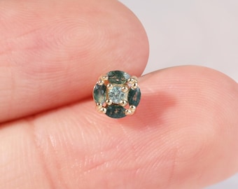 May include: A small, round stud earring with a gold setting. The earring features a cluster of small, dark green and light blue gemstones arranged in a square pattern. The earring is held between two fingers.