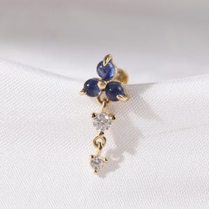 Può includere: Un orecchino dorato con un gruppo di tre pietre preziose blu scuro nella parte superiore e due pietre preziose chiare pendenti sotto. L'orecchino è posto su uno sfondo bianco strutturato.