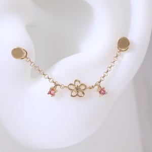 May include: A gold-colored ear cuff featuring a delicate floral design with small pink gemstones. The cuff is connected by a chain to two round studs, creating a stylish and elegant accessory.