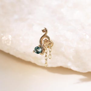 Può includere: Un delicato orecchino color oro con un design a spirale e una piccola gemma rotonda color verde acqua. Una sottile catena pende dalla parte inferiore dell'orecchino. L'orecchino è posto su uno sfondo bianco strutturato.