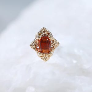 May include: A gold nose stud featuring an oval-shaped orange gemstone set in a decorative, filigree-style metal frame. The setting has a pointed, diamond-like shape with intricate metalwork details surrounding the central orange stone.