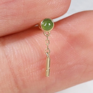 May include: A gold stud earring featuring a small, round, green cabochon stone set in a bezel. A delicate gold chain hangs from the base of the setting, ending in a slender, cylindrical gold bar charm that dangles vertically from the stud.