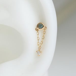 May include: A gold earring featuring a small circular stud with a blue stone, connected by two delicate gold chains to a tiny clear gemstone charm. The earring is positioned on the upper cartilage area of a white ear model for display.