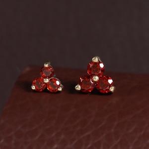 May include: A pair of stud earrings, each featuring three small, round, faceted red gemstones arranged in a triangular cluster. The stones are set in a metallic frame with visible prongs holding each individual gem securely in place.
