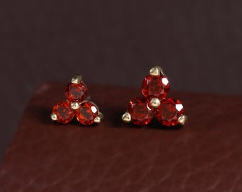 May include: A pair of stud earrings, each featuring three small, round, faceted red gemstones arranged in a triangular cluster. The stones are set in a metallic frame with visible prongs holding each individual gem securely in place.