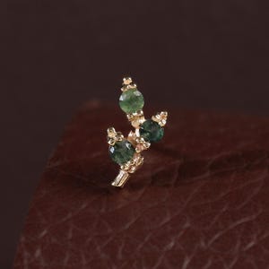 May include: A gold stud earring featuring a branch-like design with three small, round, faceted green gemstones set in gold prongs. The metal structure is delicate and slender, mimicking the appearance of a twig with tiny gold beads.