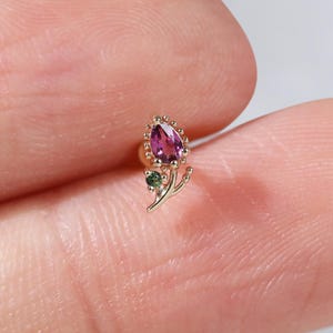 Könnte beinhalten: Ein goldener Ohrstecker in Blumenform mit einem birnenförmigen rosa Edelstein als Blütenblatt und einem kleinen runden grünen Edelstein als Blatt. Die Metallfassung weist eine feine Perlenstruktur um den rosa Stein und einen dünnen Stiel auf.