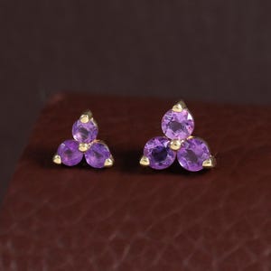 Puede incluir: Un par de pendientes de botón con un diseño de racimo de tres piedras. Cada pendiente consta de tres amatistas moradas de talla redonda engastadas en una montura de metal dorado, dispuestas en un patrón triangular similar a un trébol.