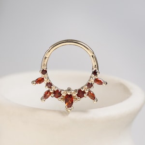 May include: A gold-toned circular hoop ring featuring a decorative segment adorned with multiple small, deep red gemstones. The stones are arranged in a fan-like pattern along the lower half of the hoop, alternating between round and marquise shapes.