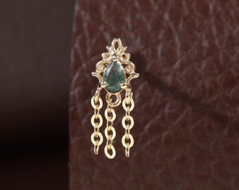 May include: A gold nose stud featuring an ornate, pointed metal frame set with a single teardrop-shaped green stone. Three delicate gold chains hang vertically from the base of the frame, creating a dangling effect on this piece of jewelry.