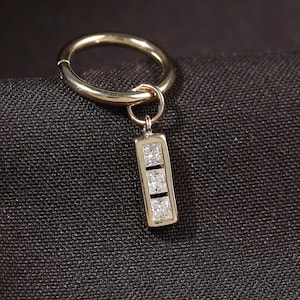 May include: A gold-colored hoop earring with a rectangular charm. The charm features three square, clear gemstones. The earring is set against a dark, textured background.