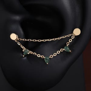 May include: A gold-toned ear cuff featuring two flat circular studs connected by two delicate chains. The lower chain is adorned with three small, teardrop-shaped green stones, creating a layered, draped effect across the ear cartilage.