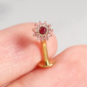 May include: A gold-tone labret stud earring featuring a flower-shaped head with a central round dark red stone surrounded by a halo of small light pink stones. The post is straight and ends in a flat circular disc base for secure placement.