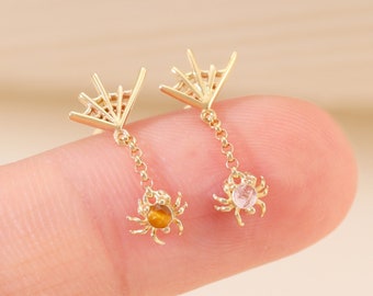 May include: A pair of gold spider earrings with dangling charms. One spider has a pink gemstone and the other has an orange gemstone.