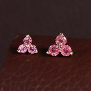 May include: A pair of stud earrings featuring a cluster of three round, faceted pink stones set in gold metal. Each earring is arranged in a triangular formation, with the stones held securely by small gold prongs at the center and edges.