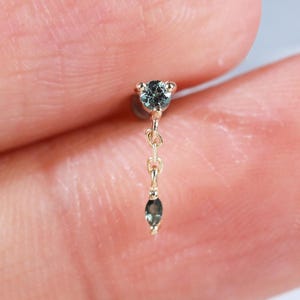 May include: A gold stud earring featuring a round, blue-grey gemstone set in a prong setting, connected by a short, delicate gold chain to a smaller, marquise-cut, blue-grey gemstone that hangs vertically from the main stud component.