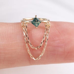 以下が含まれることがあります： 四角い、ティール色の宝石が付いたゴールドカラーのリング。リングには、3つの繊細なゴールドチェーンがぶら下がっています。白い背景に指にはめられたリング。宝石は約0.3cmです。