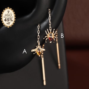 May include: Two gold-tone threader earrings hanging from a black ear display. Earring A features a bee charm with an amber-coloured stone and black accents. Earring B features a spider charm with a red stone and black accents. Both have thin chains.