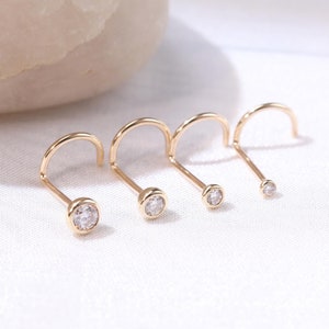 May include: Four gold nose rings with clear gemstones. The rings are arranged in a row from largest to smallest.