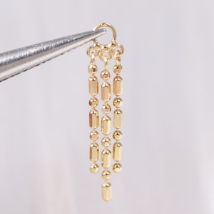 May include: A gold-colored jewelry component with a small ring at the top and three dangling strands. Each strand alternates between small round beads and elongated, rectangular beads. The component is held by a pair of silver tweezers against a white background.