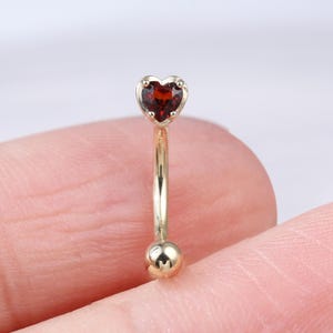 May include: A gold-coloured curved eyebrow ring featuring a heart-shaped, dark red gemstone at the top and a small ball at the bottom. The jewellery is shown against a soft, neutral background.