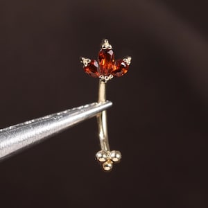 May include: A gold-coloured, curved barbell earring with a floral design. The top features three reddish-brown gemstones arranged in a petal shape. The bottom has three smaller, round, gold-coloured accents.