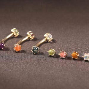 May include: Gold-toned jewelry with colorful gemstones. The image shows several curved barbell earrings and stud earrings, each featuring a different colored gemstone. The text at the bottom reads: "You can freely choose any two gemstones."