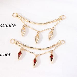 May include: Two gold-toned jewelry pieces with dangling gemstone pendants. One features clear moissanite stones, the other has deep red garnet stones. The text "Moissanite" and "Garnet" are printed on the image.