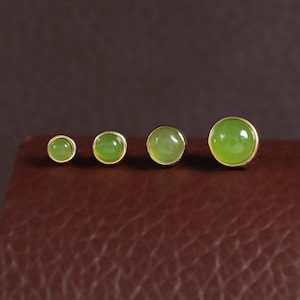 May include: Four round stud earrings arranged in a row by size, from smallest to largest. Each earring features a smooth, polished, light green cabochon stone set in a simple, thin gold-toned metal bezel frame with a flat, circular profile.