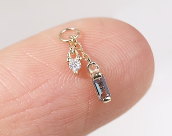 May include: A small gold charm with a dangling design. It features a small, clear, round gemstone and a rectangular, iridescent gemstone. The charm is attached to a small ring.