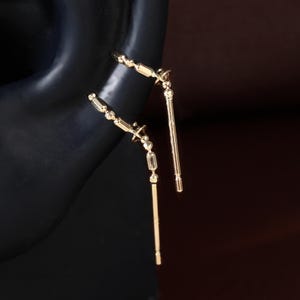 May include: Two gold-toned ear cuffs featuring a delicate chain-like design with small, cylindrical accents. Each piece consists of a thin, curved band that wraps around the ear cartilage, terminating in a straight, slender vertical post.
