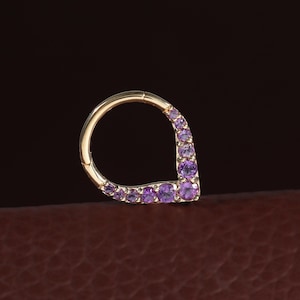 Peut inclure: Une boucle d'oreille créole en or avec un segment en forme de goutte orné d'une rangée de petites pierres précieuses rondes de couleur violette. Le bijou présente un mécanisme de fermeture à charnière et une finition métallique polie.