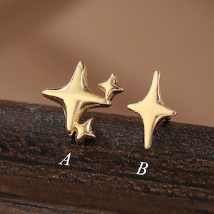 Könnte beinhalten: Zwei goldfarbene Ohrstecker aus Metall. Option A zeigt eine Anordnung aus drei vierzackigen Sternen in unterschiedlichen Größen. Option B ist ein einzelner, länglicher vierzackiger Stern. Die Buchstaben A und B sind unter dem jeweiligen Ohrstecker abgedruckt.