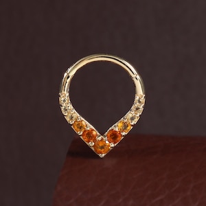 May include: A gold hoop ring featuring a V-shaped segment adorned with a gradient of round, faceted orange and yellow gemstones. The jewelry piece has a circular top section and a pointed bottom, showcasing a polished metallic finish throughout.