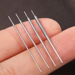 Puede incluir: Primer plano de cuatro agujas finas y plateadas. Las agujas se sostienen en una mano, con las puntas hacia arriba. Las agujas están espaciadas uniformemente y parecen ser de metal. El fondo es de color marrón oscuro borroso.