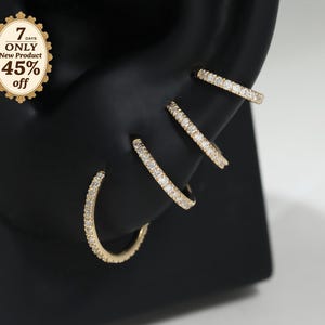 May include: Four gold-toned hoop earrings, each encrusted with small clear stones, displayed on a black ear-shaped stand. A decorative label on the stand reads 7 DAYS ONLY New Product 45% off. The hoops vary in size and are arranged in a row.