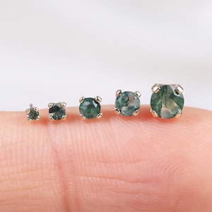 Może przedstawiać: Pięć złotych kolczyków sztyftów z okrągłymi, zielonymi kamieniami z mchu agatowego o różnych rozmiarach. Kolczyki są prezentowane na palcu na miękkim, białym tle. Kamienie mają unikalny, naturalny wzór.