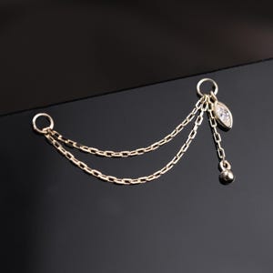 May include: A gold ear cuff featuring two fine chain drops. A small, clear marquise-cut gemstone and a tiny ball pendant hang from the chains. The ear cuff is designed to be worn on the ear.
