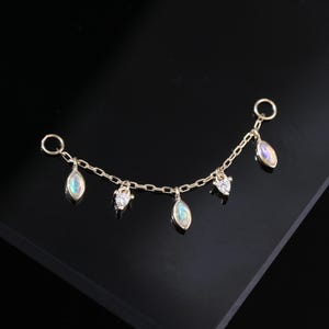 Puede incluir: Una delicada cadena dorada con colgantes. La cadena presenta piedras opalescentes en forma de lágrima y pequeñas gemas brillantes en forma de corazón. La joya está diseñada para ser usada como una cadena para la oreja o como joyería corporal.