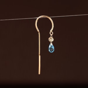 May include: A gold threader earring featuring a curved wire, a thin chain with a straight bar end, and a dangling charm. The charm consists of a small, textured gold bead positioned above a faceted, pear-shaped light blue gemstone drop.
